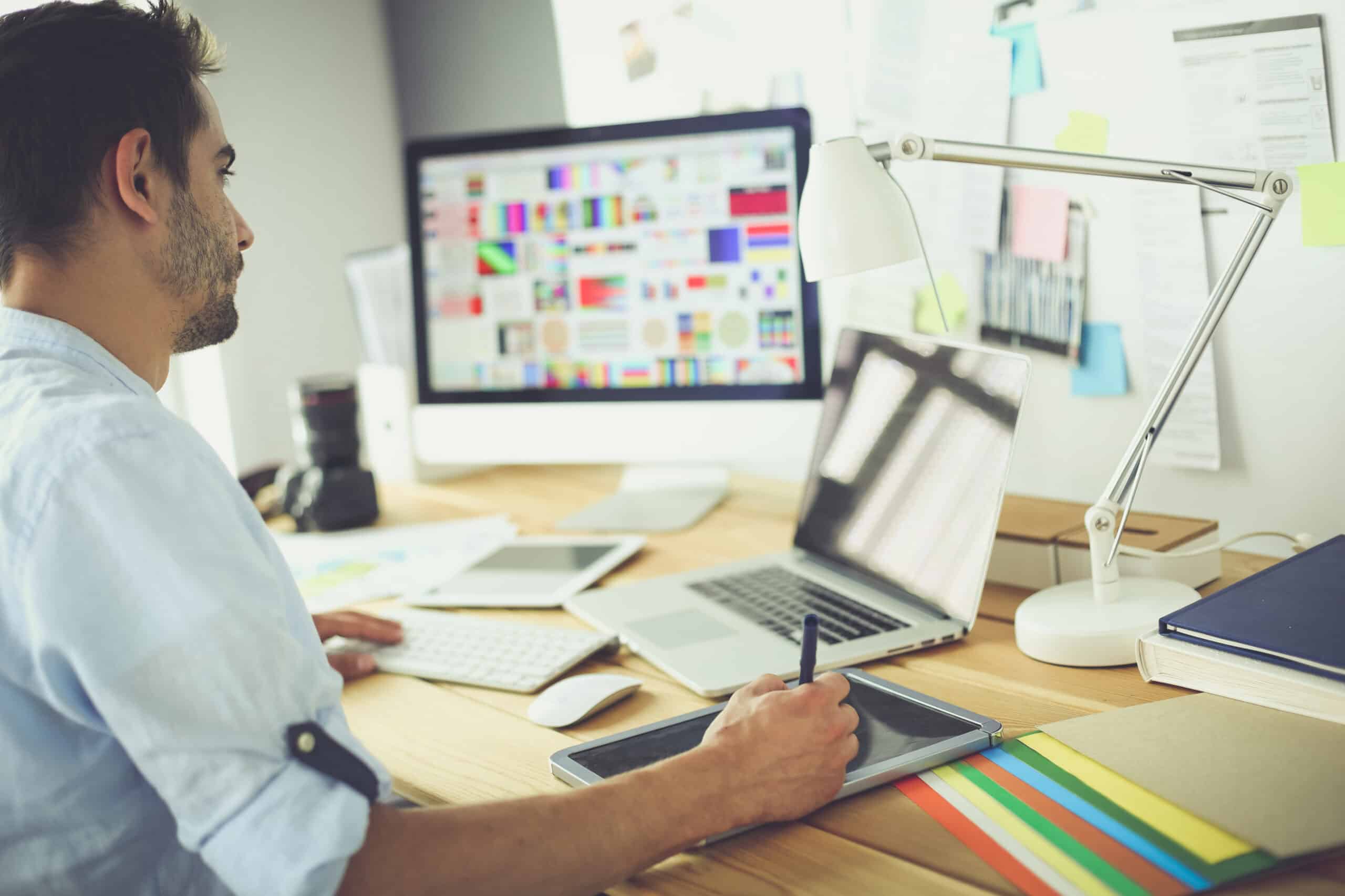 A man working at a desk with multiple screens displaying colorful designs. He is using a digital pen and tablet while looking at a computer.