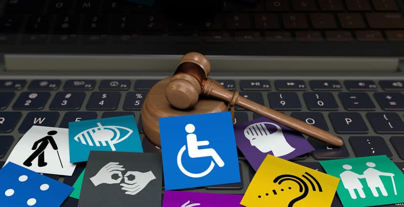 A wooden gavel resting on a laptop keyboard surrounded by colorful accessibility icons, including symbols for wheelchair access, sign language, visual and hearing impairments, and elderly care.