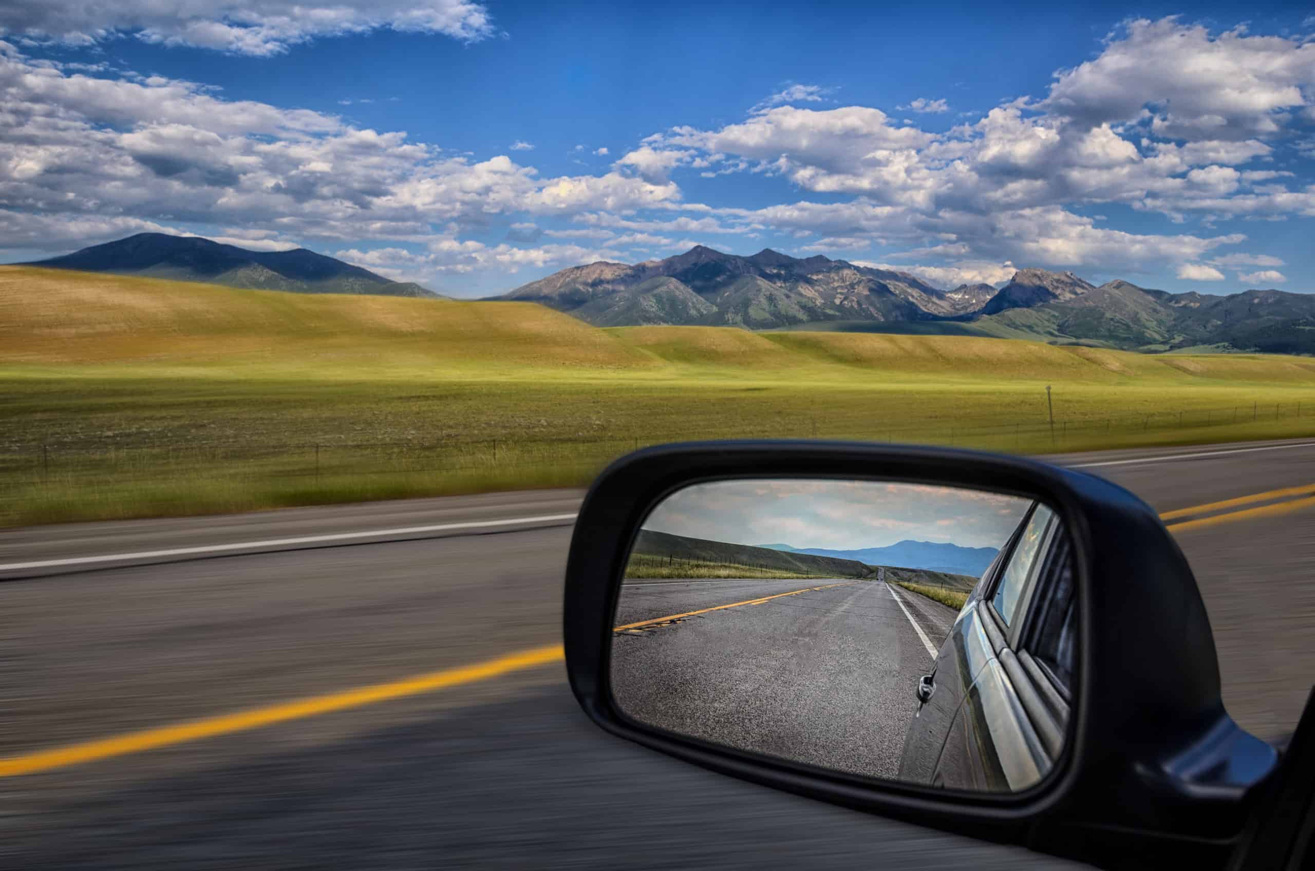 Side mirror of a vehicle with clear view of the road behind as the road ahead while larger view outside of mirror of grassy hills and mountains speed past in the distance