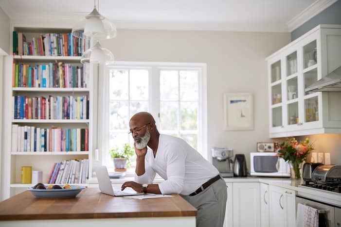 Older person using a laptop at counter in home with books and kitchen appliances in the background