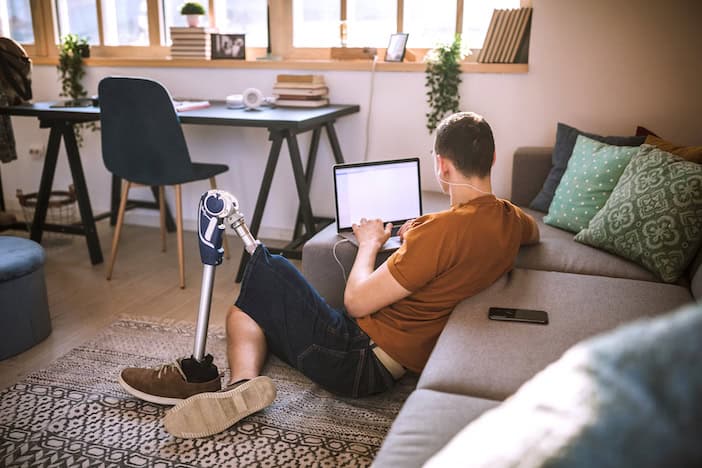 User with prosthetic left leg using laptop seated on the floor of a house on a sunny day