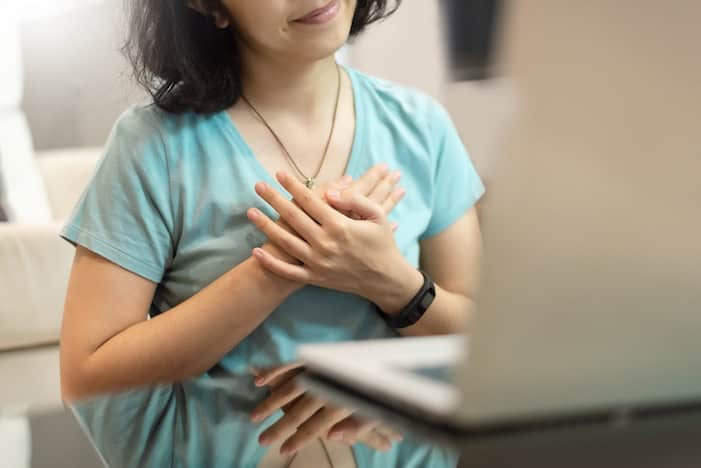 Young woman in blue shirt with sincere smile showing grateful gesture with touching on her chest with hands. Asian girl feeling thankful people with using laptop computer and internet technology.